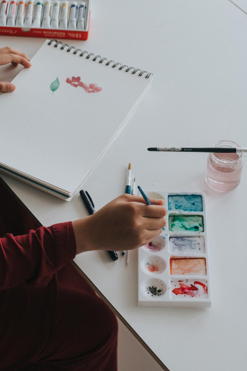 personne en train de peindre avec un pinceau sur une feuille blanche avec une palette de couleurs à droite et des tubes de peinture à gauche